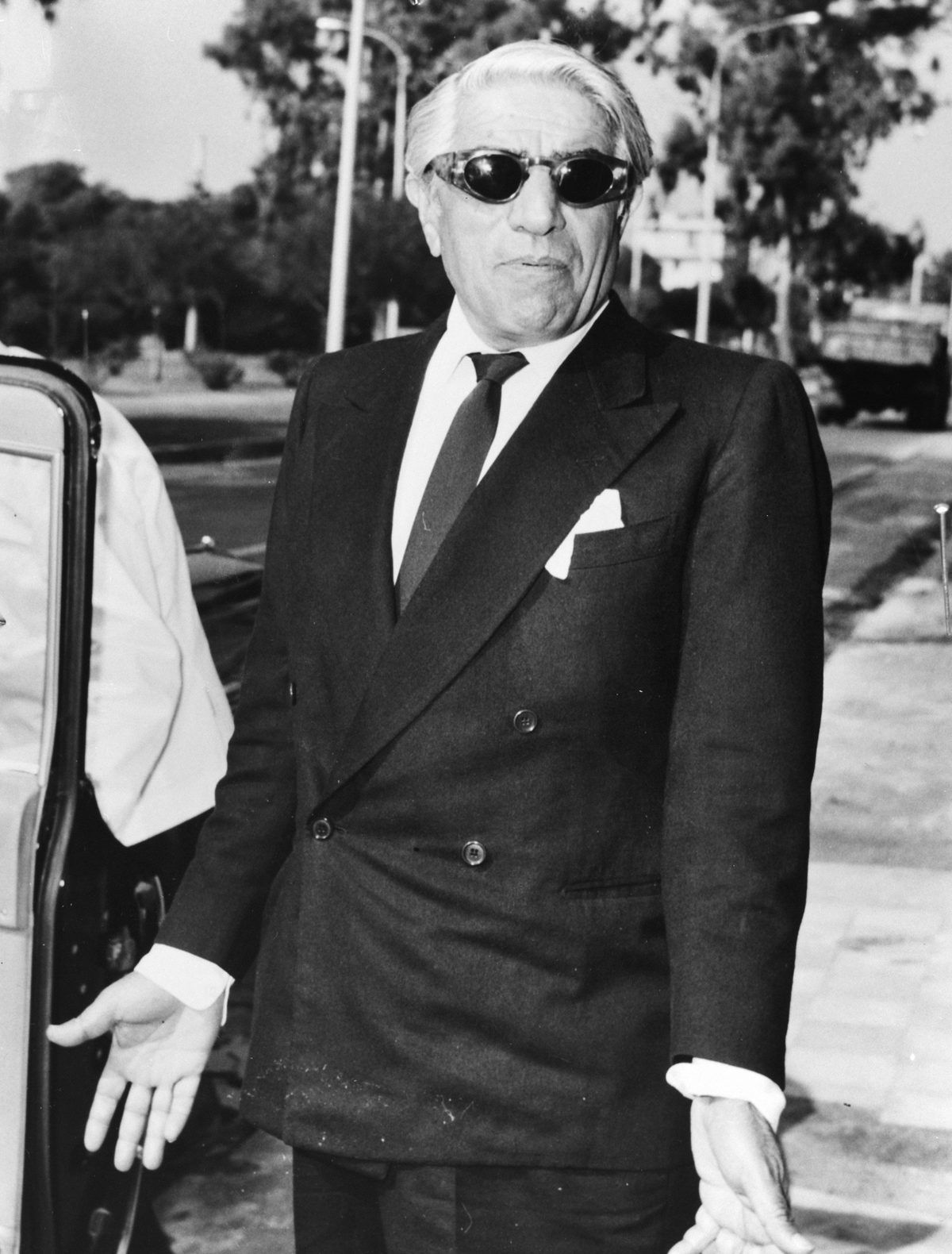 Aristote Onassis en costume croisé et lunettes noires, devant une voiture, en 1968